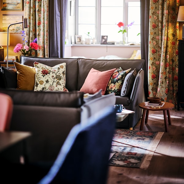 A BACKSÄLEN 3-seat sofa with a lot of different cushions lying on it, sits near a window in a living room.