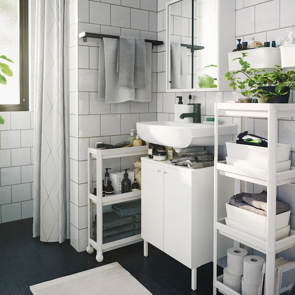 A bathroom VESKEN trolley and shelving unit with towels, plants and beauty products, on either side of the wash-stand.