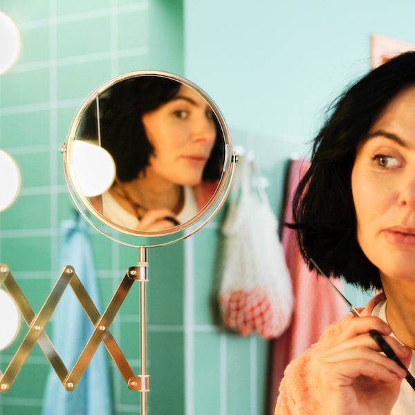 A bathroom with green tiles, a woman is looking into a FRÄCK mirror while cutting her own hair.