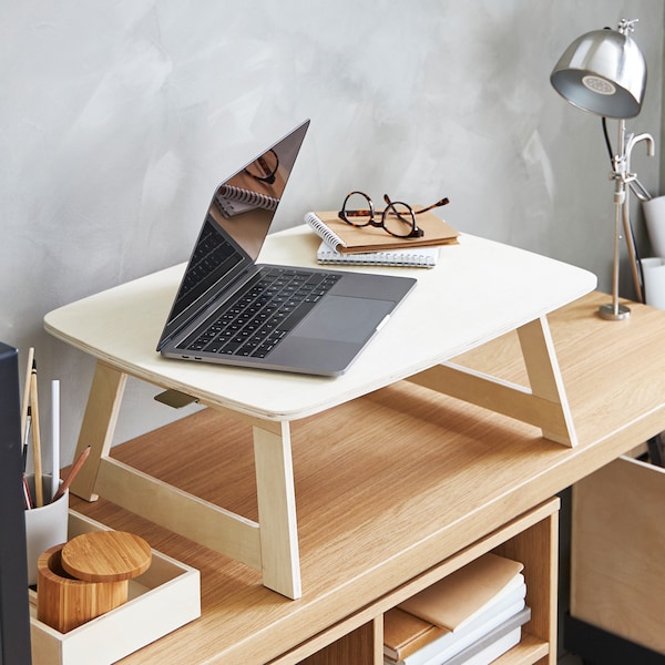 A birch laptop stand with a laptop and spectacles on it on top of an oak veneer console table with a clamp table lamp.