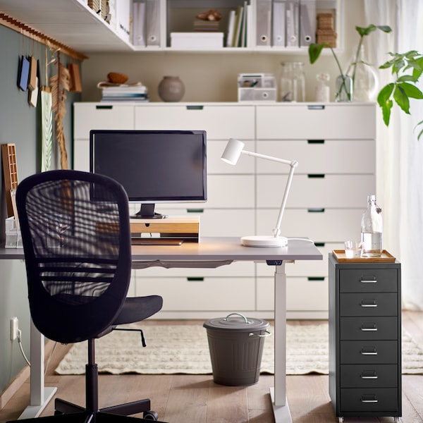 A black FLINTAN office chair and HELMER drawer unit by a grey RODULF sit/stand desk with a work lamp and computer screen.