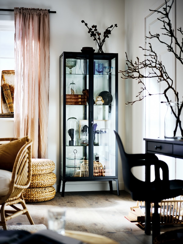 A black MILSBO cabinet with glass doors filled with an assortment of objects, in the corner of a bright living room.