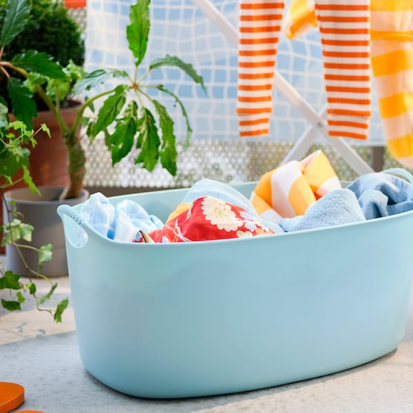 A blue TORKIS flexi laundry basket filled with laundry in front of a drying rack and a potted plant.