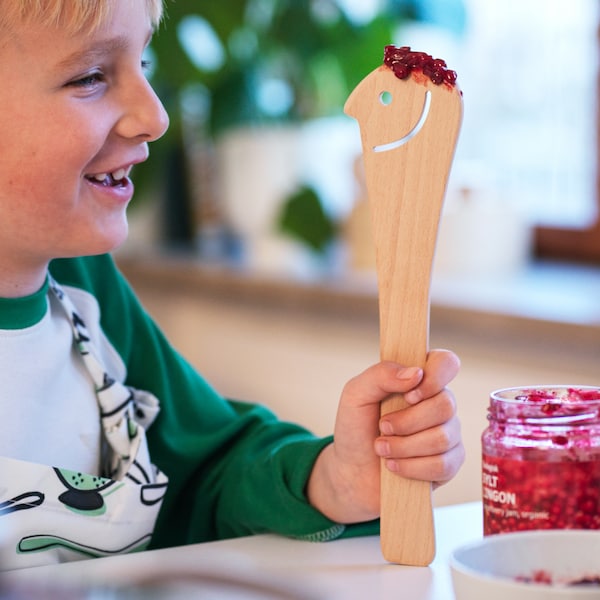 A boy has a wooden UPPLIVAD spoon smeared with SYLT LINGON spread on a kitchen counter, the spoon has a smiley face cut-out.