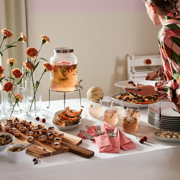 A buffet table with different dishes and a VARDAGEN glass jar filled with a peach-colored drink being poured into glasses.