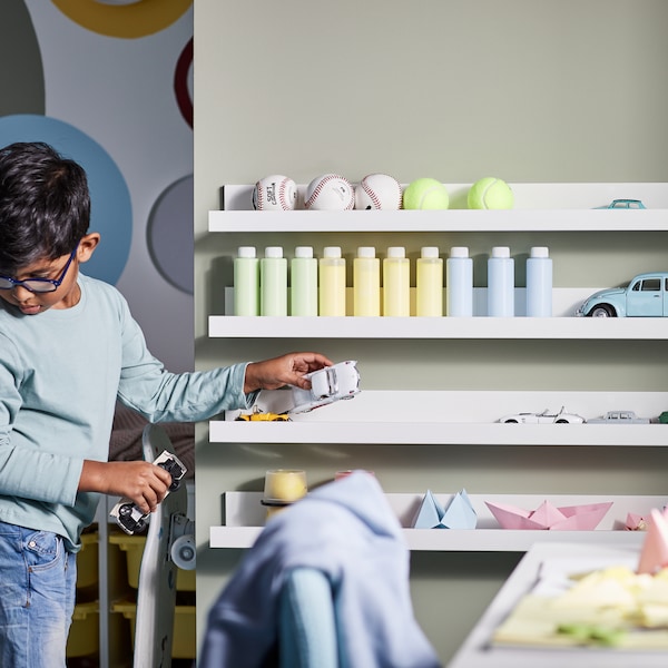 A child playing with toy cars in a bedroom with white MOSSLANDA picture ledges on the wall holding paper models and bottles.