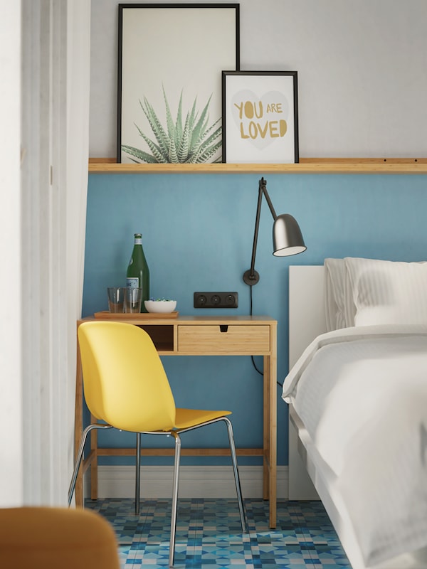 A corner of a white bed next to a window with NORDKISA dressing table, a black work/wall lamp and a yellow chair.