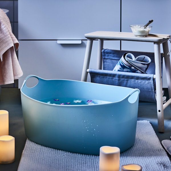 A cozy bathroom set-up with a blue TORKIS laundry basket filled with water on the floor and LED candles placed around.