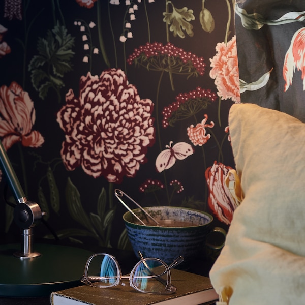 A cup and a book with a pair of glasses on it sit on a bedside table near a pillow, in front of a wall with a floral pattern.