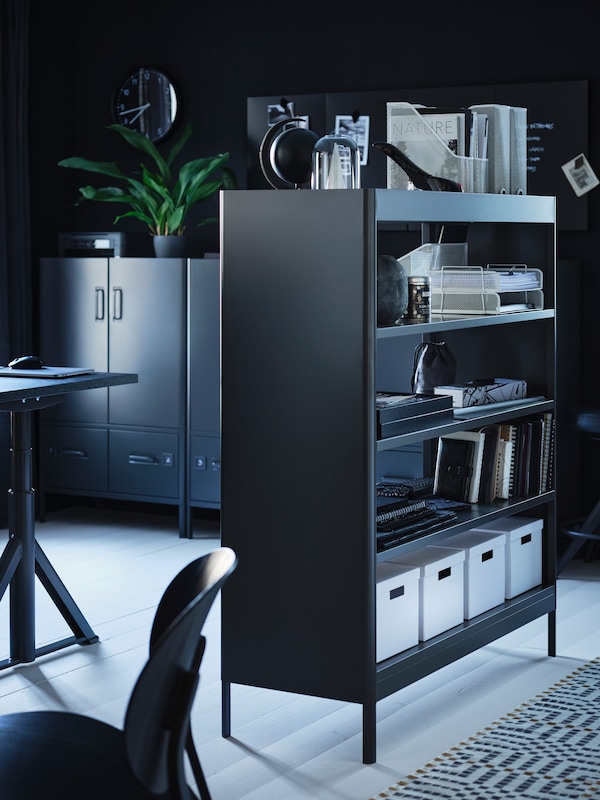 A dark grey IDÅSEN shelving unit used as a room divider in a home workspace, holding storage boxes, books and other items.