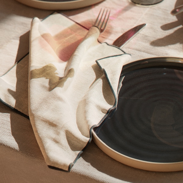 A dark grey OMBONAD plate sits on a table with an OMBONAD tablecloth beside an OMBONAD napkin on top of a knife and fork.