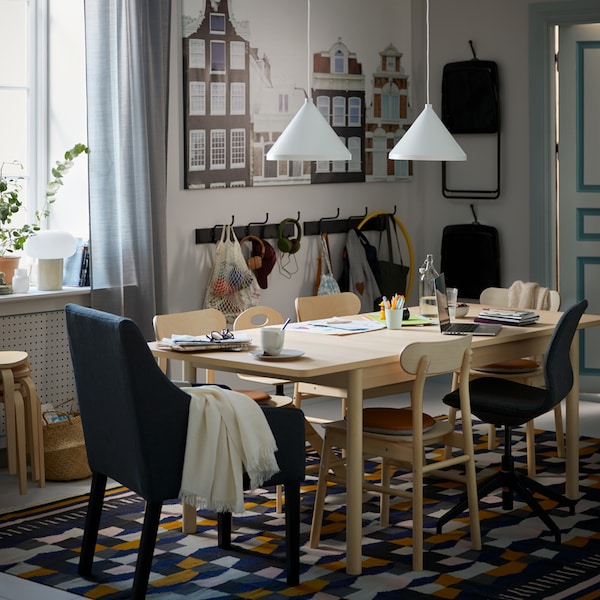 A dining area with RÖNNINGE birch table and chairs on a multicolour TÅRBÄK rug, with white NÄVLINGE pendant lamps above.