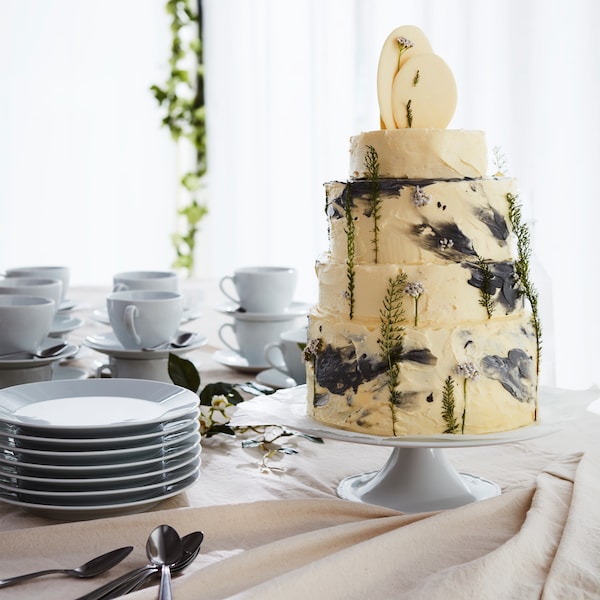 A festive table with a tall cream-colored cake with decorations placed next to stacked white plates and cups.