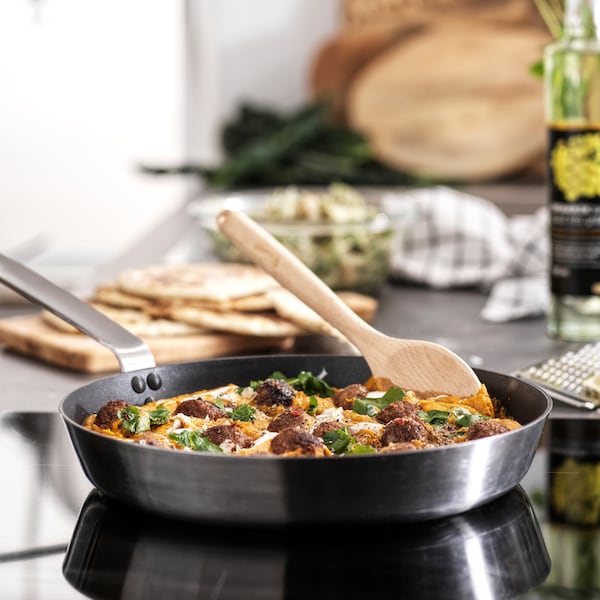 A frying pan on a cooktop filled with food, with a wooden spoon in it. A bottle of oil and a chopping board is behind it.