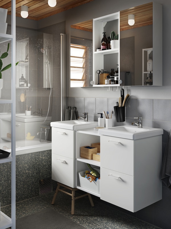 A grey bathroom with two wash-basins in white, two wall cabinets with mirror doors and bath towels and a bath mat in grey.