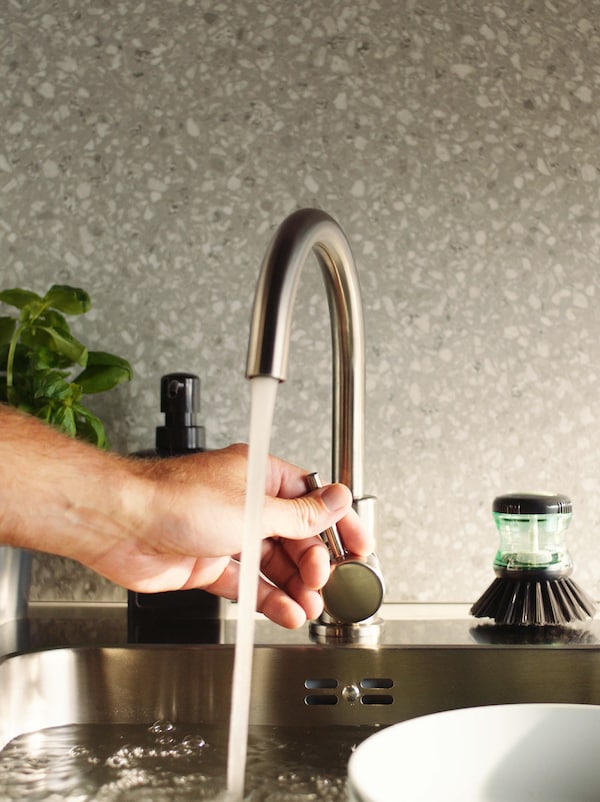 A hand controlling the flow of water from an arched mixer tap into a sink, in front of a grey wall panel with a plant beside it.