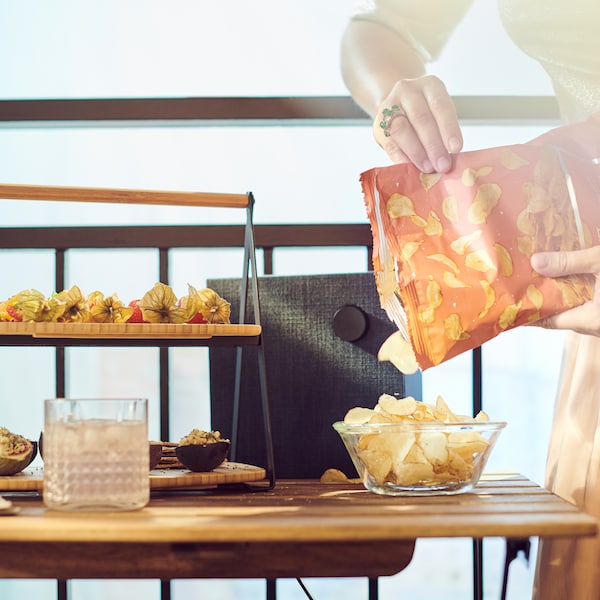 A hand pouring potato crisps into a bowl on a table that also holds a serving tray filled with lots of different snacks.