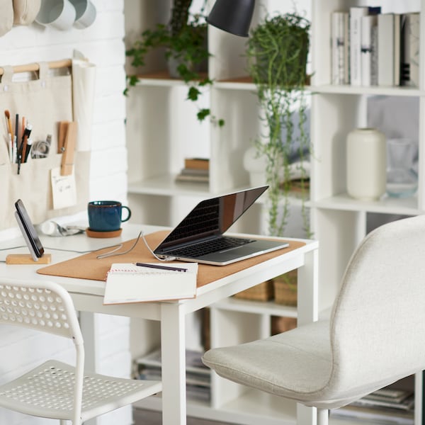 A laptop on a white table with one white and one beige office chair, beside a white shelving unit with books, and storage.