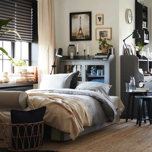 A light beige sofa-bed made as a bed with grey bedding. Behind is a grey glass-door cabinet and a jute rug on the floor.