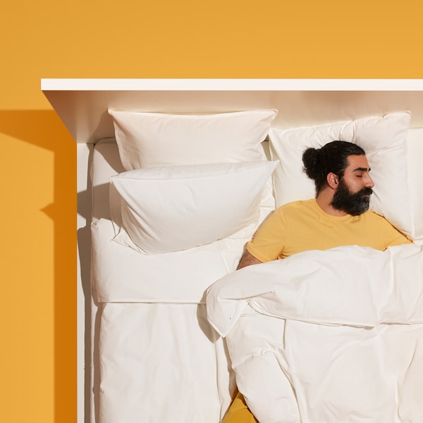 A man with a beard and yellow pajamas lies sleeping in a white bed with white bedding which stands on a yellow floor.