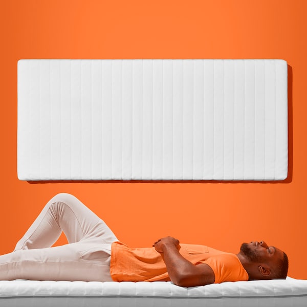 A man with a beard lies sleeping on a mattress. Behind him, another mattress is attached to an orange wall.