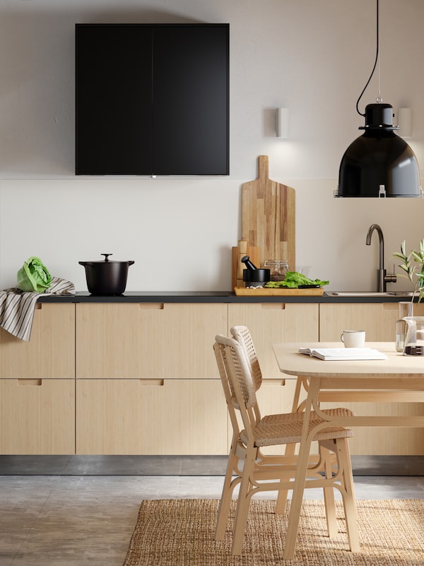 A minimalist kitchen with light bamboo drawer fronts, a light bamboo dining table and two chairs with a lamp hanging above.