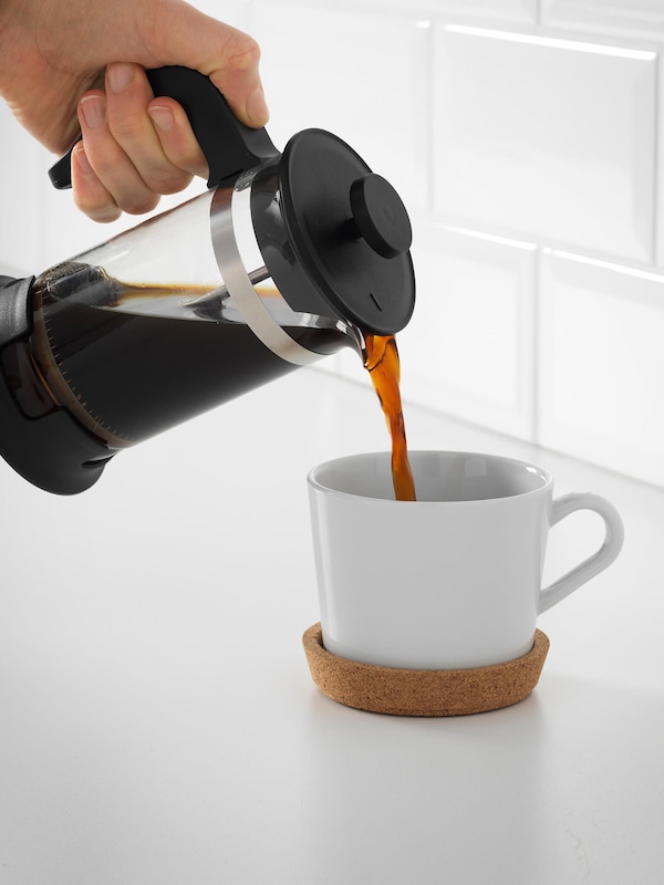 A person pours coffee from an espresso maker into a coffee mug with a wooden bottom. 