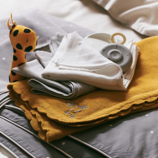 A pile of baby textiles including a dark yellow SOLGUL blanket and a LENAST crib quilt, with a baby monitor on top.