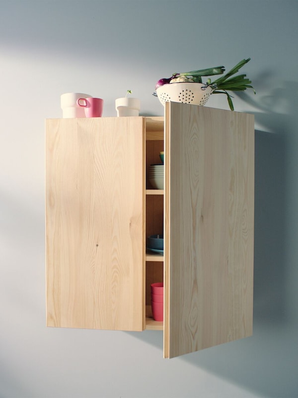 A pine, IVAR cabinet mounted on a wall with one door partially open, objects inside and on top.