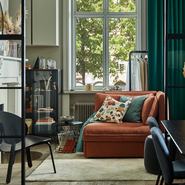 A rust sofa bed in the middle of a living room with grey walls, green curtains, a glass door cabinet and a beige rug.