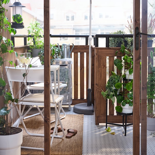 A small, leafy balcony with a white table, glasses on the table, and two chairs on a rug beside a BRAMSÖN parasol base.