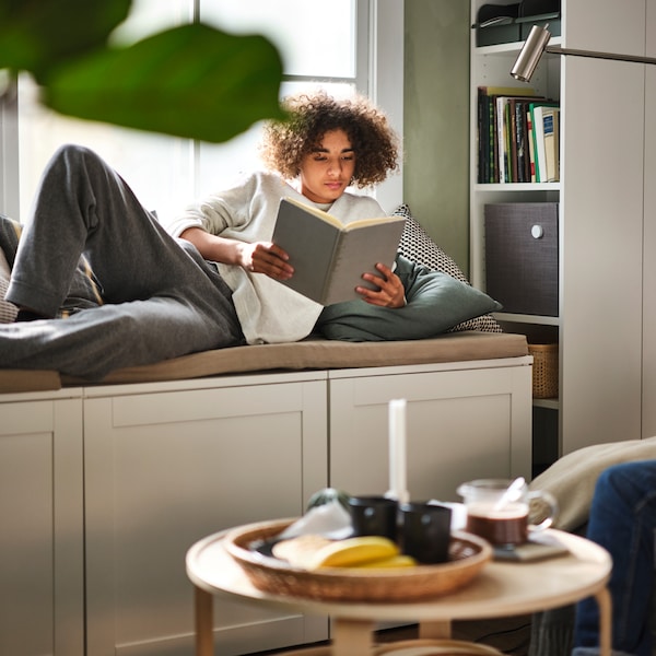 A teen reading while lying on a storage bench in front of a white BILLY bookcase, filled with books and a speaker.