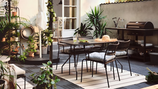 A terrace with a VIHOLMEN table surrounded by four LÄCKÖ chairs. An outdoor kitchen with charcoal barbecue is by the wall.