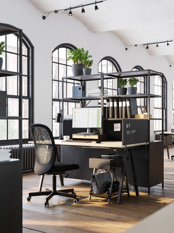 A TROTTEN desk and black office chair by open shelves with a privacy board at one side of the desk and a storage unit below.