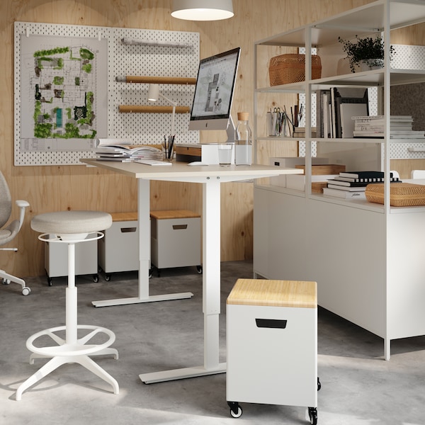 A TROTTEN sit/stand desk with a LIDKULLEN active sit/stand support in front in a bright, white studio with open shelving.