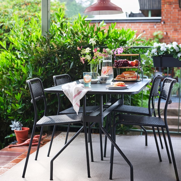 A VIHOLMEN table and four chairs on a terrace with a jug of water and a serving tray holding sandwiches and fruit on top.