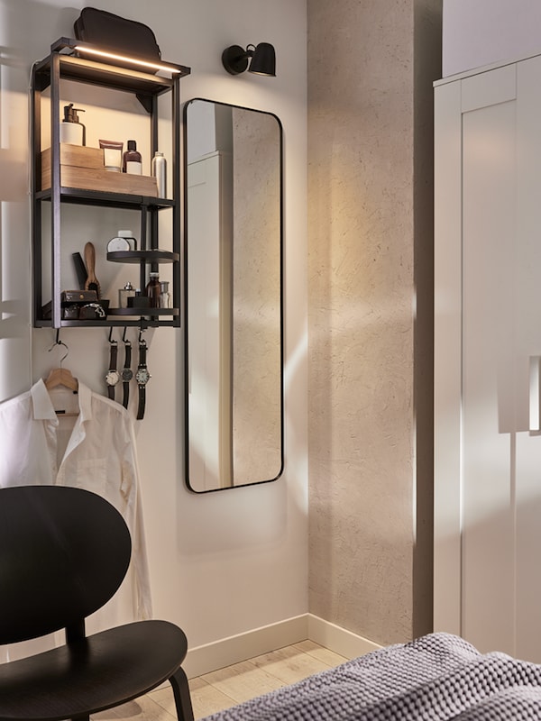 A wall with a mirror, wall lamp and anthracite wall frame with shelves with toiletries, watches and a shirt hanging from it.