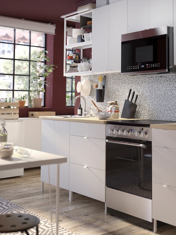 A white ENHET kitchen combination in a deep red room with a desk in front of it. A microwave is above the CIRKULERA range.