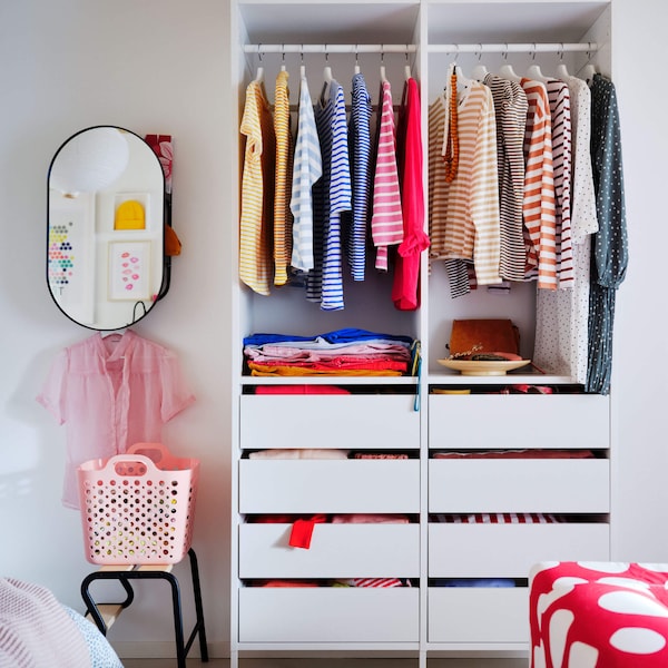 A white storage unit with hanging clothes on clothe srails in a room with a mirror mounted on the wall.