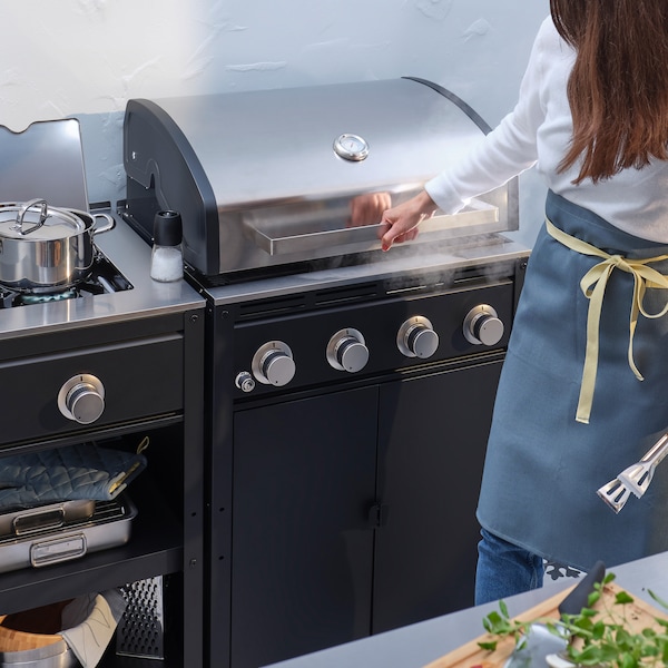 A woman holding barbecue tongs prepares to open the lid on the gas barbecue grill of a GRILLSKÄR outdoor kitchen.