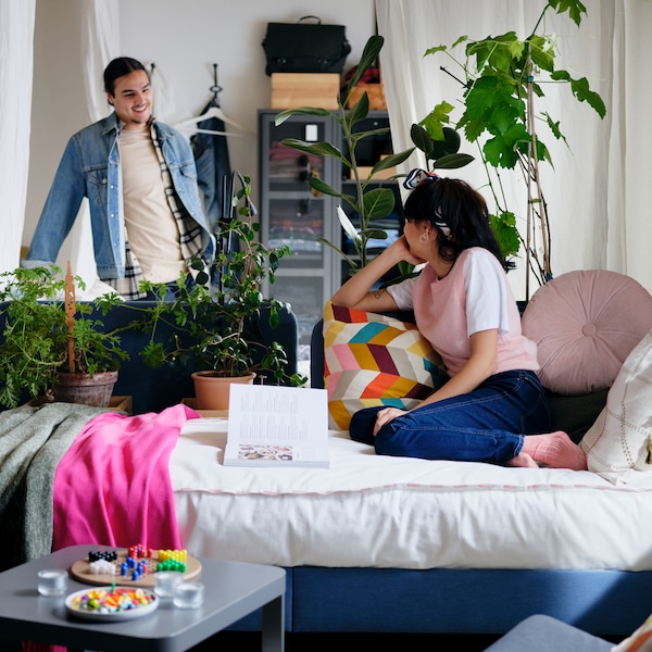 A woman on a blue BLÅKULLEN bed with various colourful cushions and throws, with a man standing behind some plants.