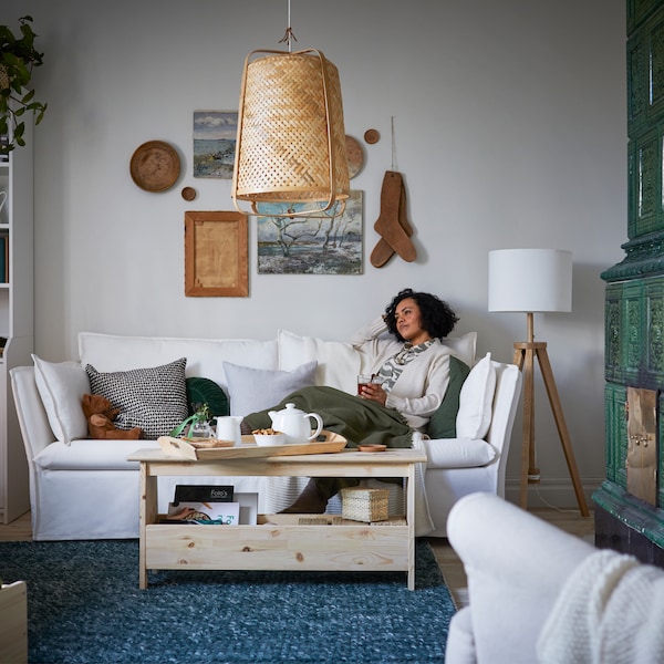 A woman relaxes on a white BACKSÄLEN sofa, covered in a throw, with a cup of tea.