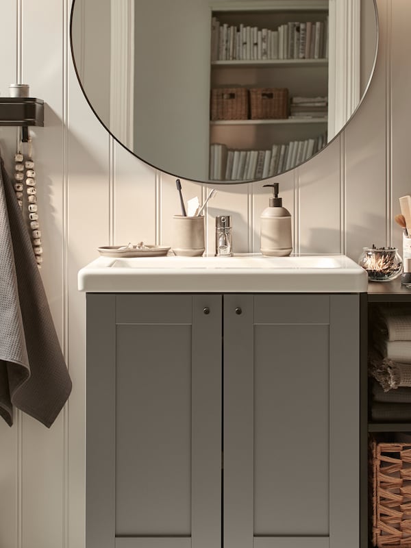 An ENHET/TVÄLLEN wash-basin cabinet with two doors by an open shelving unit, against a panelled wall with a mirror on it.