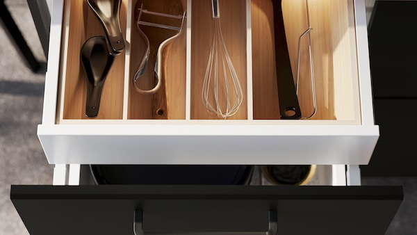 An open kitchen drawer with a black front, revealing kitchen utensils inside an UPPDATERA cutlery tray in light bamboo.