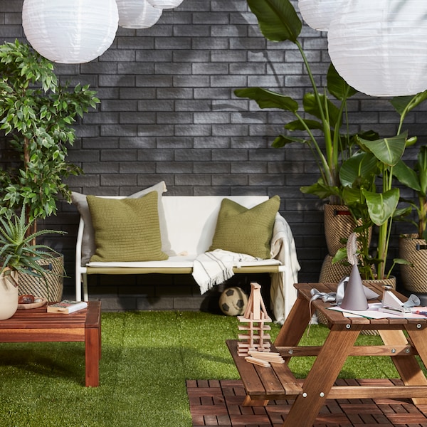 An outdoor area with a cosy seating area, lush plants, SOLVINDEN LED solar-powered pendant lamps against a grey brick wall.