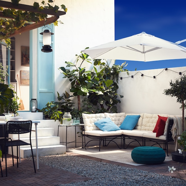 An outdoor patio with a modular sofa with white cushions, a white parasol, a blue-green pouffe and plenty of plants.