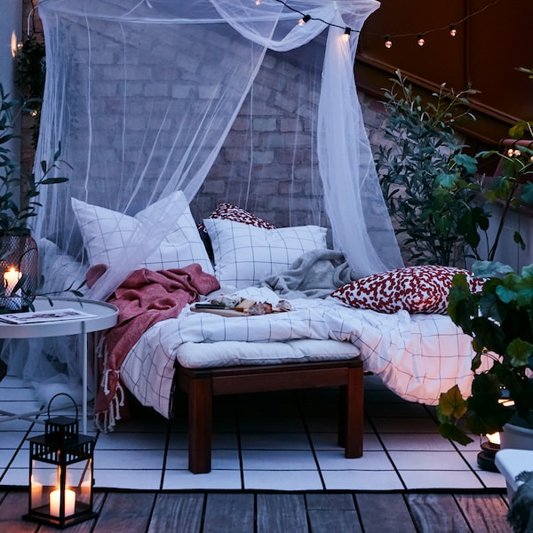 An outdoor terrace at dusk with a cozy area made up of a lounging bed, SOLIG net, rug, lanterns and outdoor lighting chains.