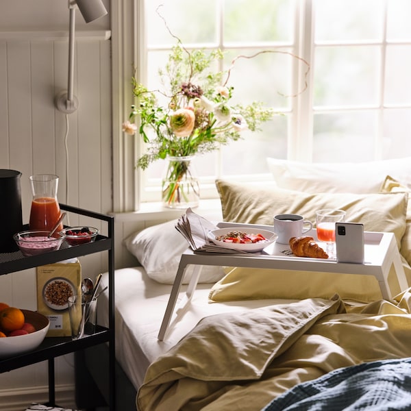 Breakfast served on a white KLIPSK bed tray on a bed made with ÄNGSLILJA bed linen, with more food on a trolley.