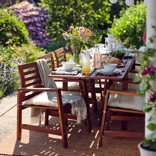 Eight ÄPPLARÖ wooden chairs and a table laid for a meal on top of a rug on a large, leafy terrace.