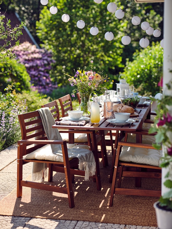 Eight ÄPPLARÖ wooden chairs and a table laid for a meal on top of a rug on a leafy terrace with an LED lighting chain above.